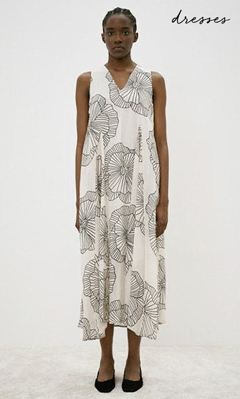 A person wearing a sleeveless, mid-calf white dress with a black abstract floral pattern stands against a plain light background. The word “dresses” is written in cursive in the top right corner. Fearrington Village