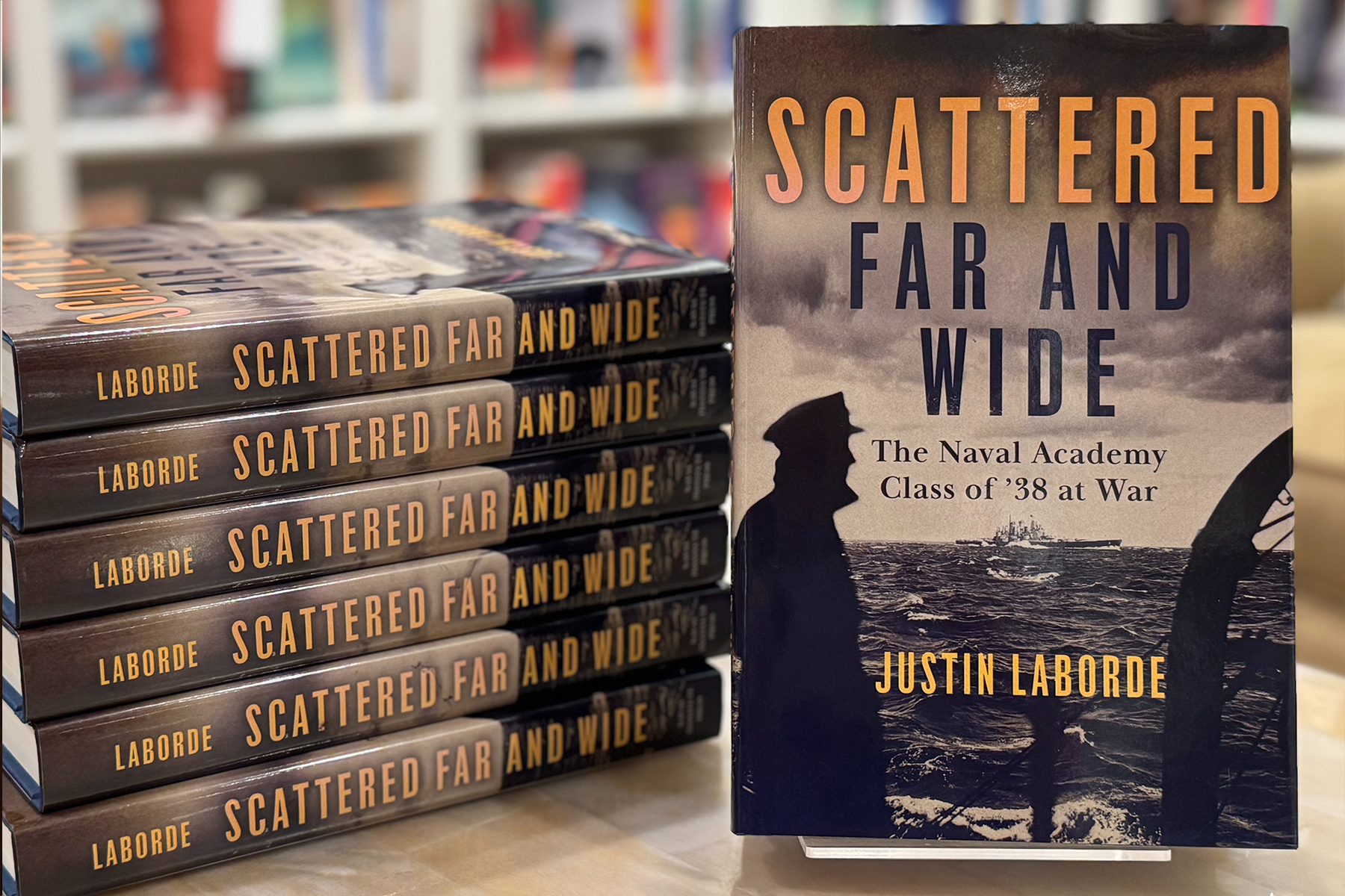 A stack of books titled "Scattered Far and Wide" by Justin Laborde is displayed on a table, with one book standing upright to show its cover featuring a silhouette of a naval officer and a cloudy sky. Fearrington Village