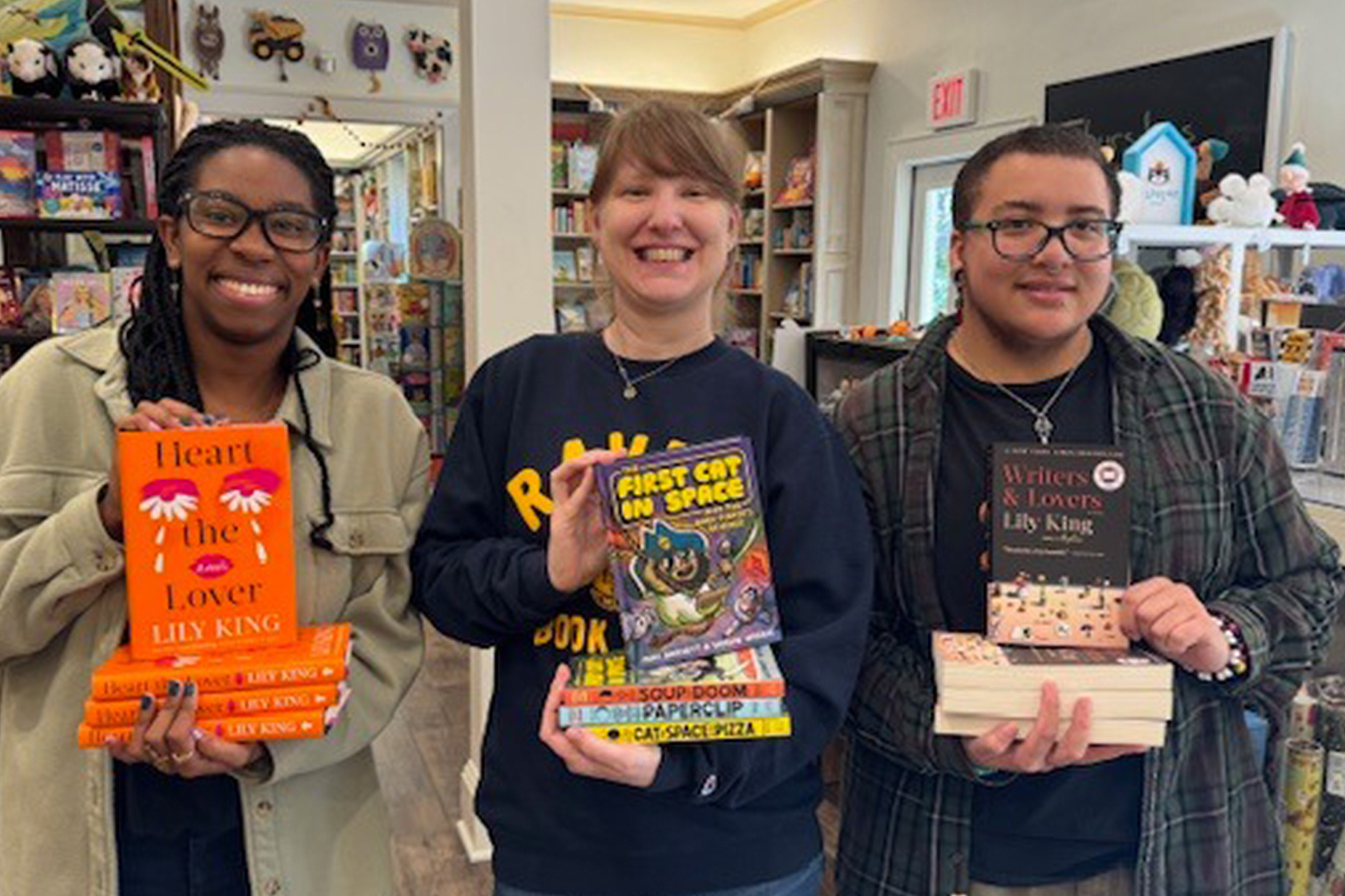 Three people stand indoors holding stacks of books and smiling. The person on the left holds copies of "Writers & Lovers," the middle person holds "First Cat in Space," and the right person holds various books. Bookstore shelves are in the background. Fearrington Village