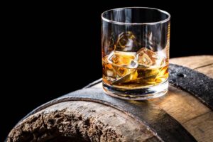 A glass of whiskey with ice sits on top of a wooden barrel, against a black background. Fearrington Village