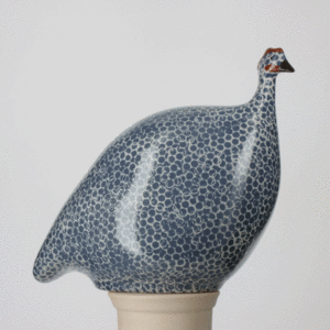A ceramic sculpture of a guinea fowl with a rounded body, covered in blue and white spotted patterns, and a small head with brown and white details, set on a neutral base against a plain background. Fearrington Village