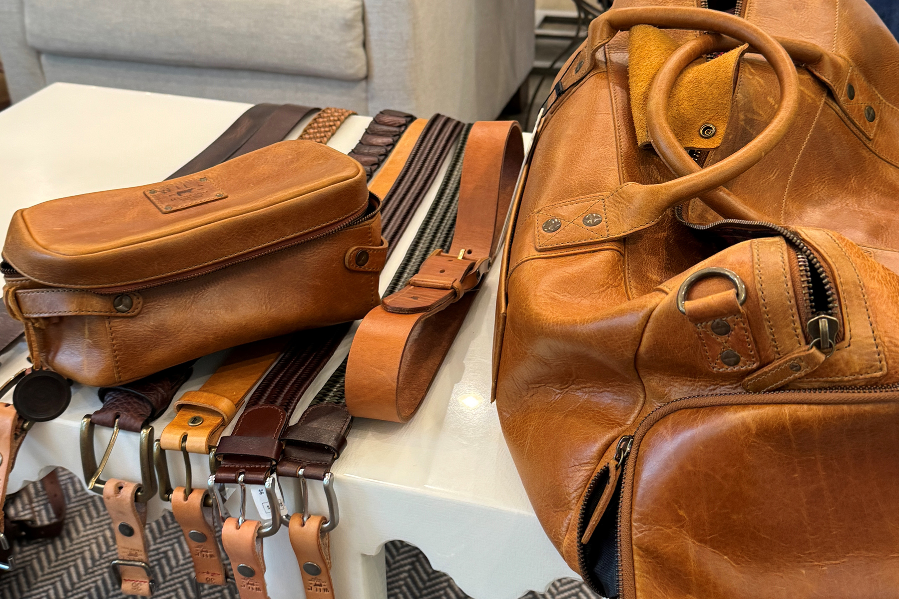 A display of tan and brown leather accessories, including a duffel bag, a smaller pouch, and several belts, arranged neatly on a white table with a gray upholstered chair in the background. Fearrington Village