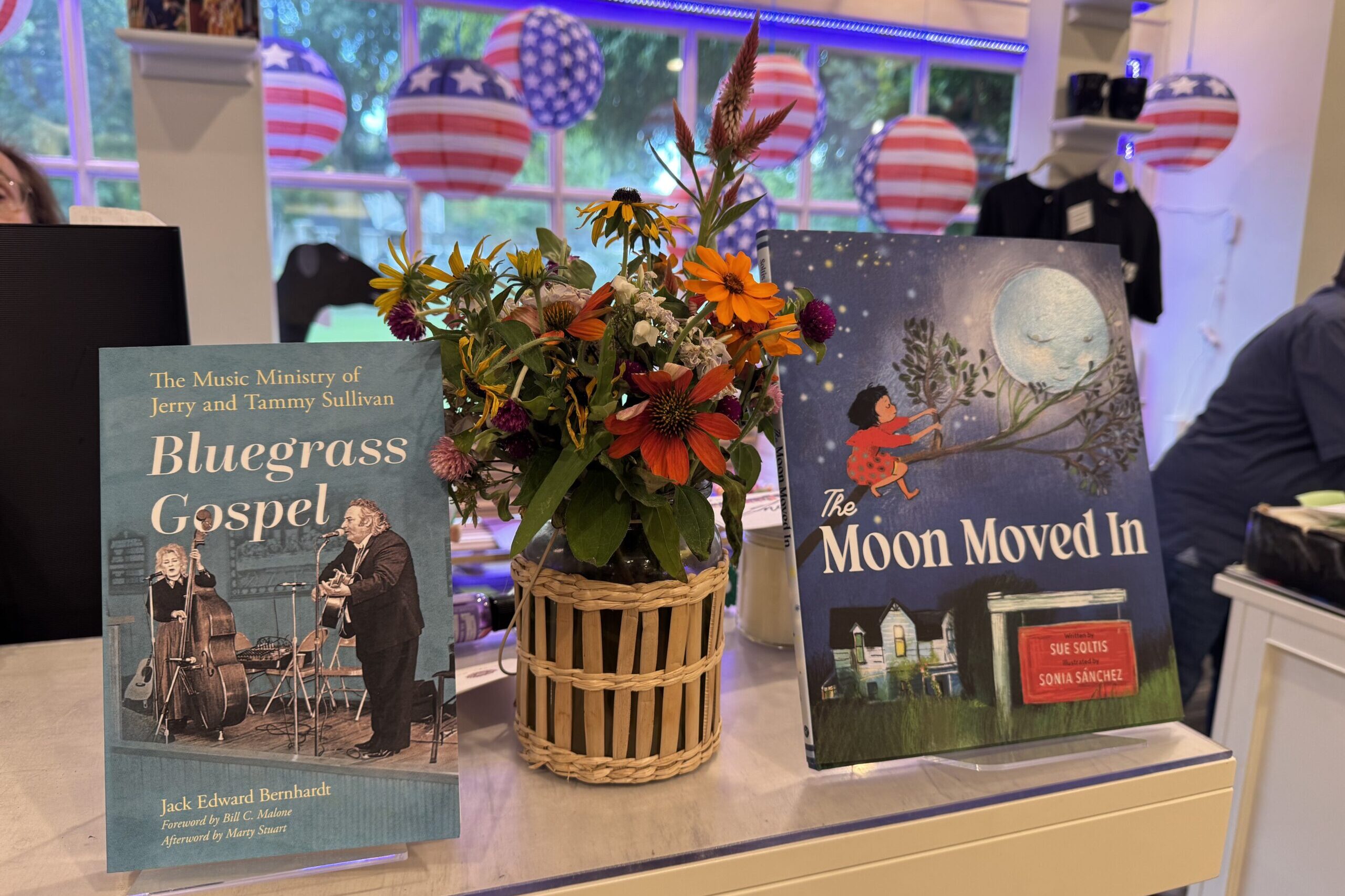 Gentle Reader - July 22, 2025 1 A vase of colorful flowers sits on a counter between two books: "Bluegrass Gospel" with musicians on the cover, and "The Moon Moved In" with a girl reaching for the moon. Patriotic American flag decorations are in the background. Fearrington Village