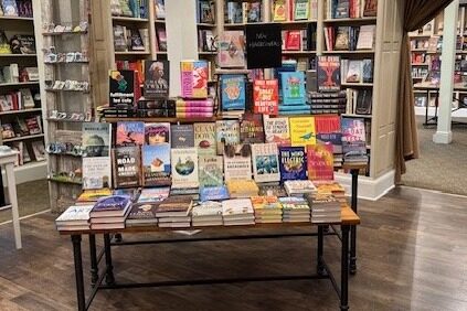 A table in a bookstore displays a variety of neatly arranged books with colorful covers. Bookshelves filled with more books line the walls in the background, creating a cozy and inviting atmosphere. Fearrington Village
