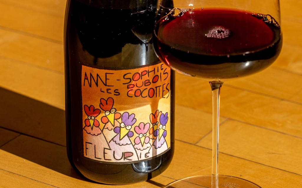 A bottle of Anne-Sophie Dubois Fleurie wine with a colorful label featuring cartoon chickens, next to a glass of red wine on a wooden surface in warm sunlight. Fearrington Village
