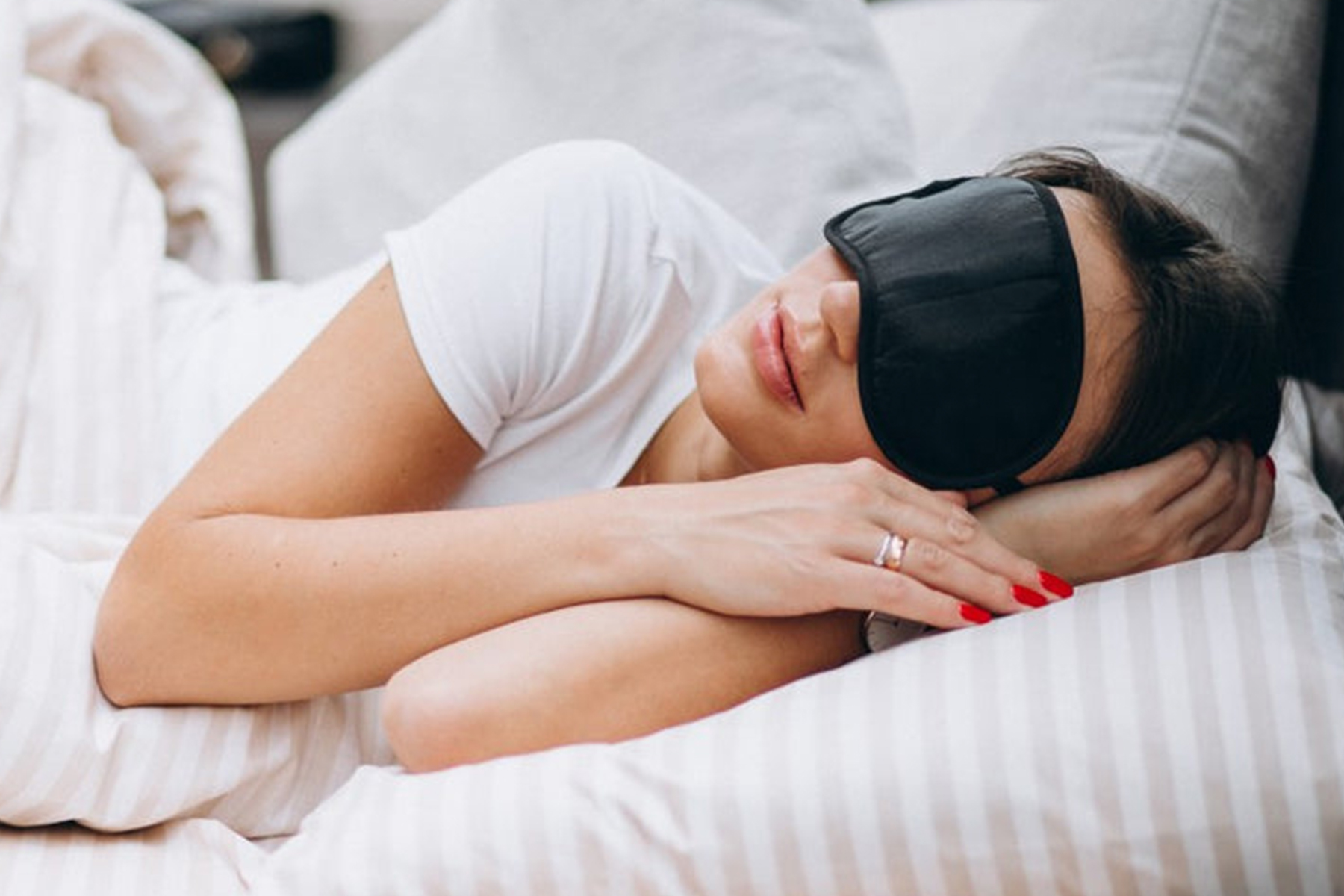 A woman wearing a black sleep mask and white shirt is lying on her side in bed, resting her head on a striped pillow, with her hands tucked under her cheek. Fearrington Village