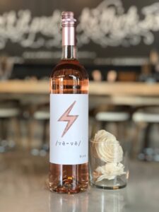 A bottle of rosé wine with a lightning bolt label stands next to a glass holding white decorative flowers on a reflective surface, with a blurred bar and stools in the background. Fearrington Village