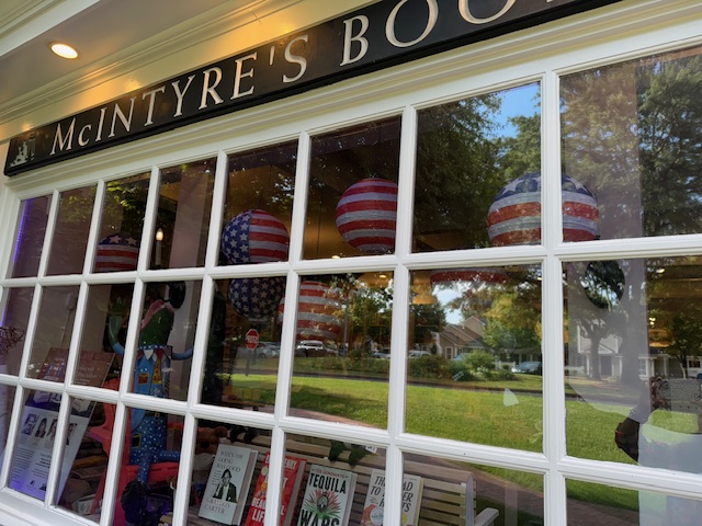 The exterior of McIntyre’s Books features large windows displaying books and American flag-themed paper lanterns, with trees and houses reflected in the glass. Fearrington Village