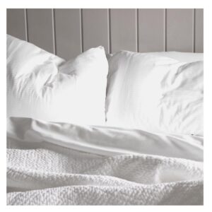 Two white pillows and a white textured blanket on an unmade bed, with a light-colored wood panel wall in the background. Soft daylight highlights the clean, cozy, and inviting bedding. Fearrington Village