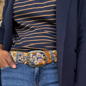 A person wearing a navy cardigan, navy and tan striped shirt, blue jeans, and a colorful embroidered belt with a large silver buckle. Their left hand is in their jeans pocket. Fearrington Village