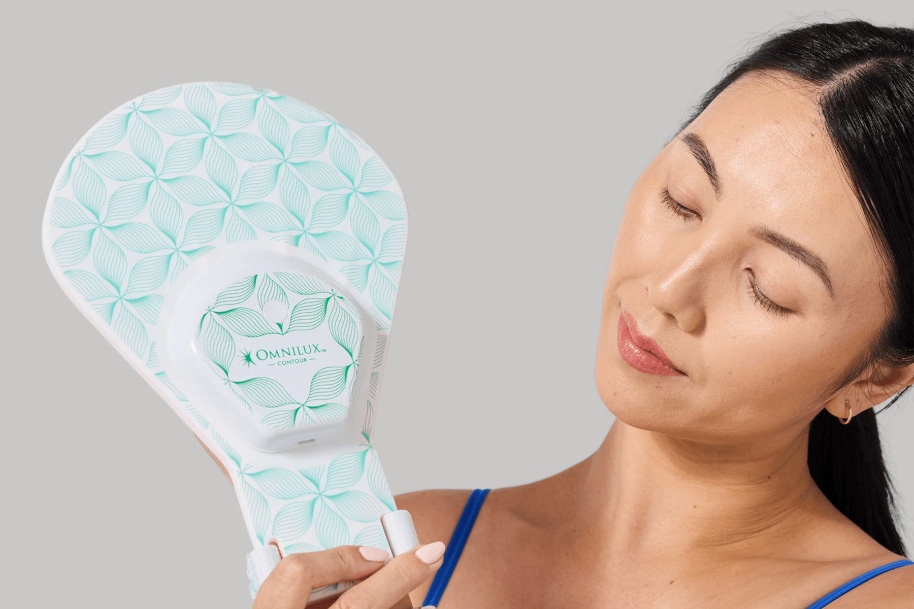 A woman with closed eyes holds a patterned Omnilux Contour device near her face, against a plain light gray background. Fearrington Village