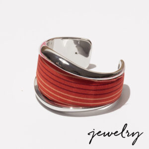 A silver cuff bracelet with a wide, red and orange striped fabric inlay is displayed on a white background. The word "jewelry" is written in black script in the lower right corner. Fearrington Village