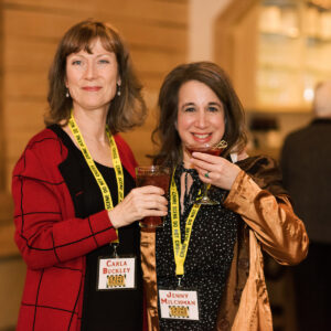 Two women, both wearing name tags and holding drinks, smile at the camera at the Mystery Book Festival. The woman on the left wears a red jacket; the one on the right wears a gold satin jacket. People are blurred in the background. Fearrington Village