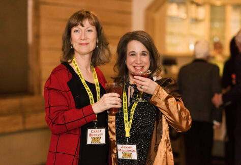 Two women, both wearing name tags and holding drinks, smile at the camera at the Mystery Book Festival. The woman on the left wears a red jacket; the one on the right wears a gold satin jacket. People are blurred in the background. Fearrington Village