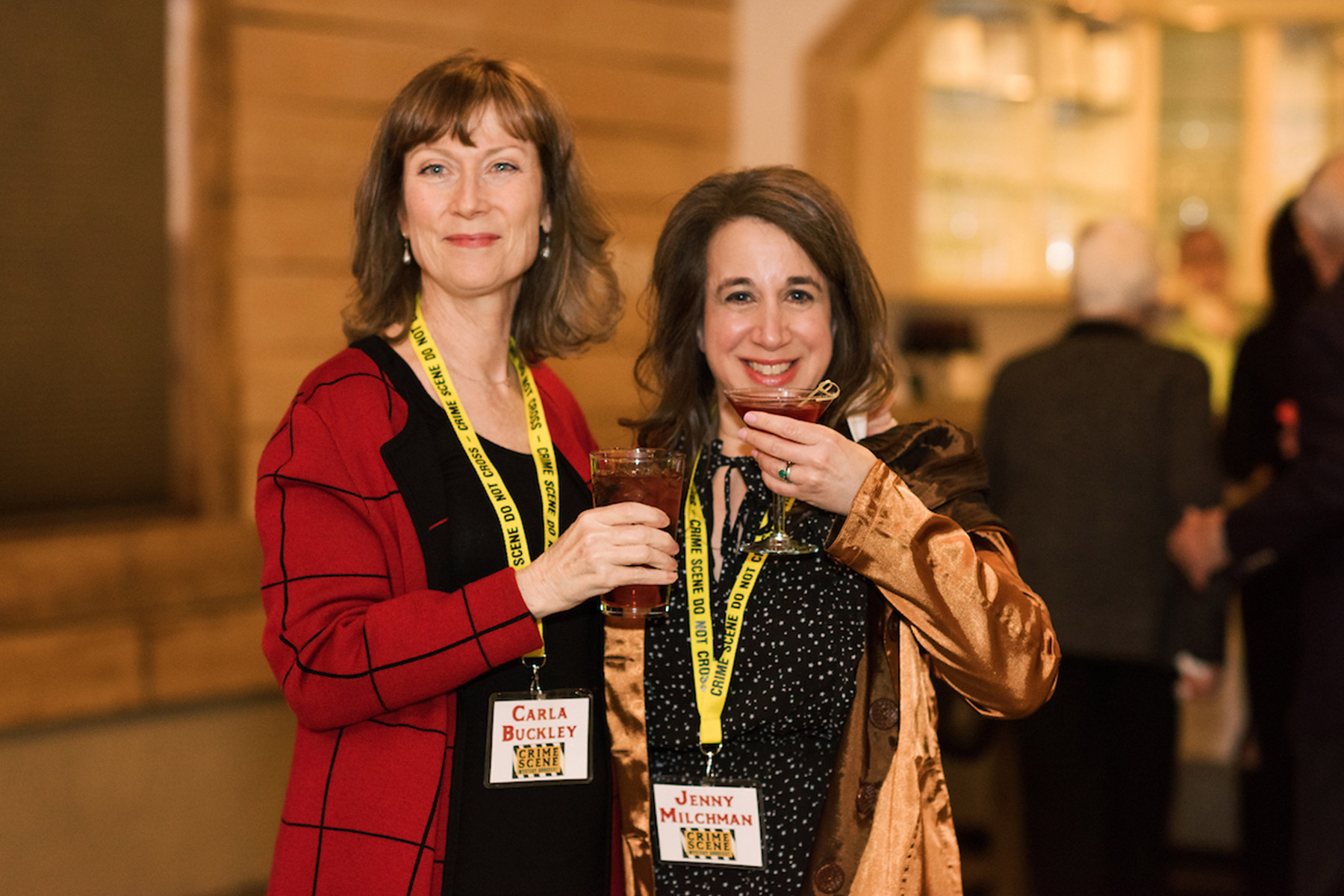 CrimeScene Killer Cocktail 2026 27 Two women, both wearing name tags and holding drinks, smile at the camera at the Mystery Book Festival. The woman on the left wears a red jacket; the one on the right wears a gold satin jacket. People are blurred in the background. Fearrington Village