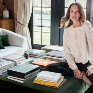 A woman in a white sweater and black pants sits cross-legged on a green ottoman covered with stacked books, next to a white couch with green pillows, in a cozy room with large windows and wooden walls. Fearrington Village