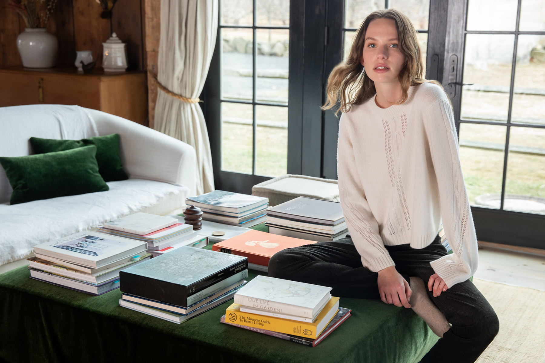 Dovecote Fall 2025 Trunk Show - Kinross 8 A woman in a white sweater and black pants sits cross-legged on a green ottoman covered with stacked books, next to a white couch with green pillows, in a cozy room with large windows and wooden walls. Fearrington Village