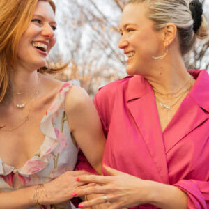 Two women smiling and laughing while walking arm in arm outdoors. One wears a floral dress, the other a bright pink jacket, both with layered necklaces. Trees without leaves are visible in the background. Fearrington Village