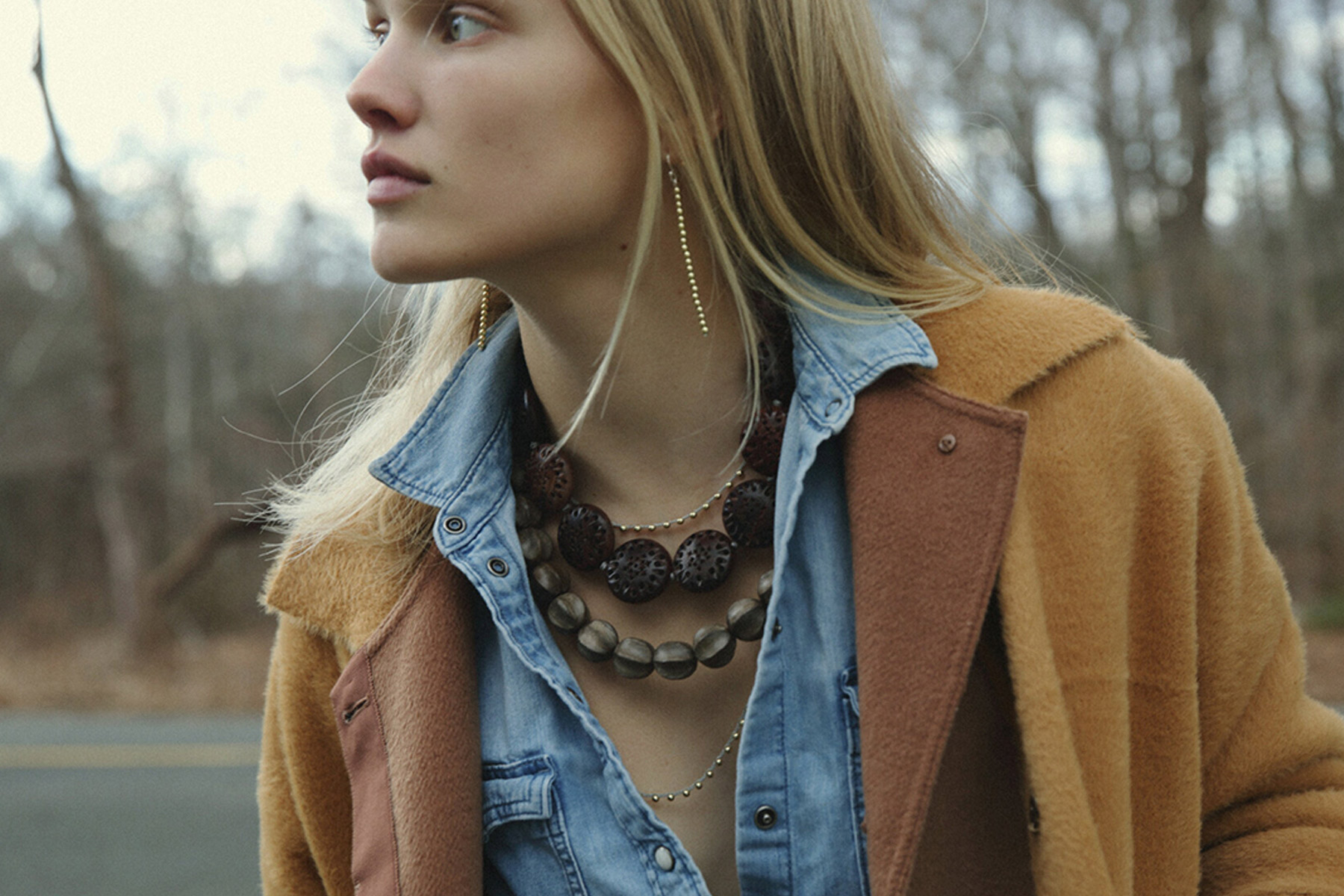 A woman with long blonde hair wears a brown coat over a denim shirt, accessorized with layered beaded necklaces and a gold earring, standing outdoors with leafless trees in the background. Fearrington Village