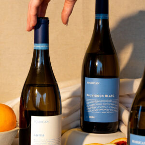 A hand reaches for a blue-capped wine bottle among three bottles labeled "Massican," next to a white bowl filled with oranges and a peach half on a beige cloth. Fearrington Village