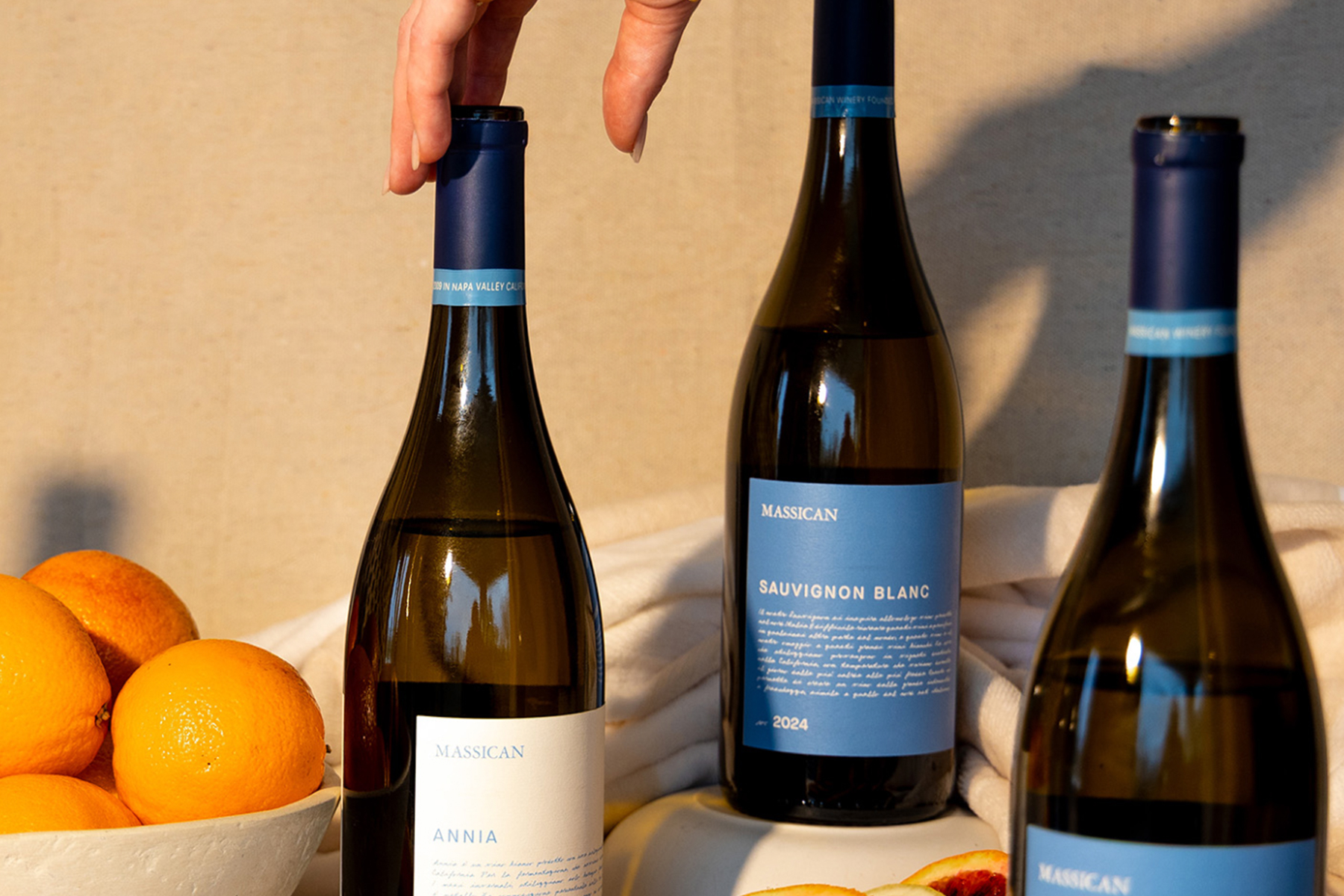 A hand reaches for a blue-capped wine bottle among three bottles labeled "Massican," next to a white bowl filled with oranges and a peach half on a beige cloth. Fearrington Village