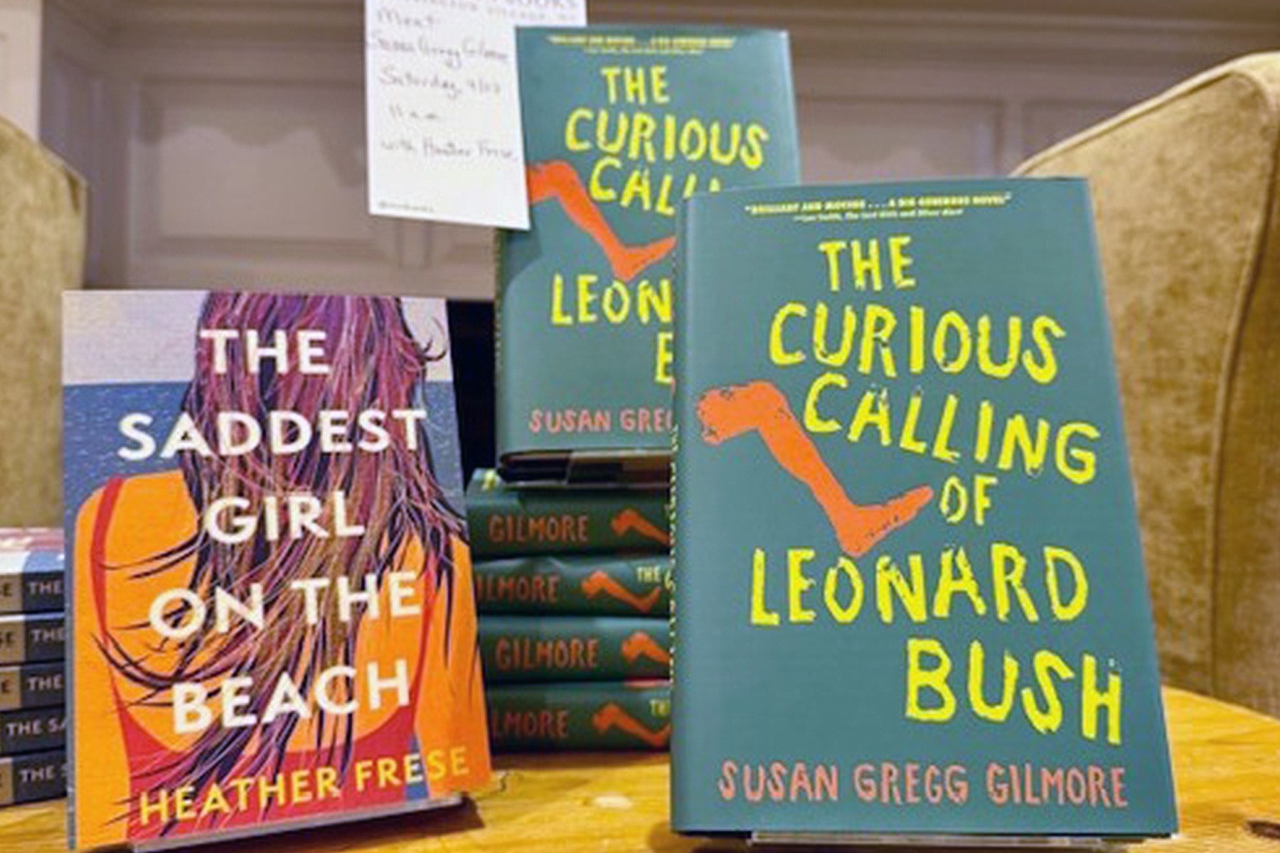 Books displayed on a table, including "The Saddest Girl on the Beach" by Heather Frese and multiple copies of "The Curious Calling of Leonard Bush" by Susan Gregg Gilmore. Fearrington Village