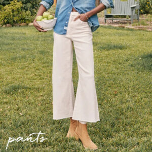 Person standing on grass wearing wide-leg cream pants, tan ankle boots, and a blue denim shirt, holding a bowl of green apples. Handwritten text "pants" appears in the lower left corner. Fearrington Village