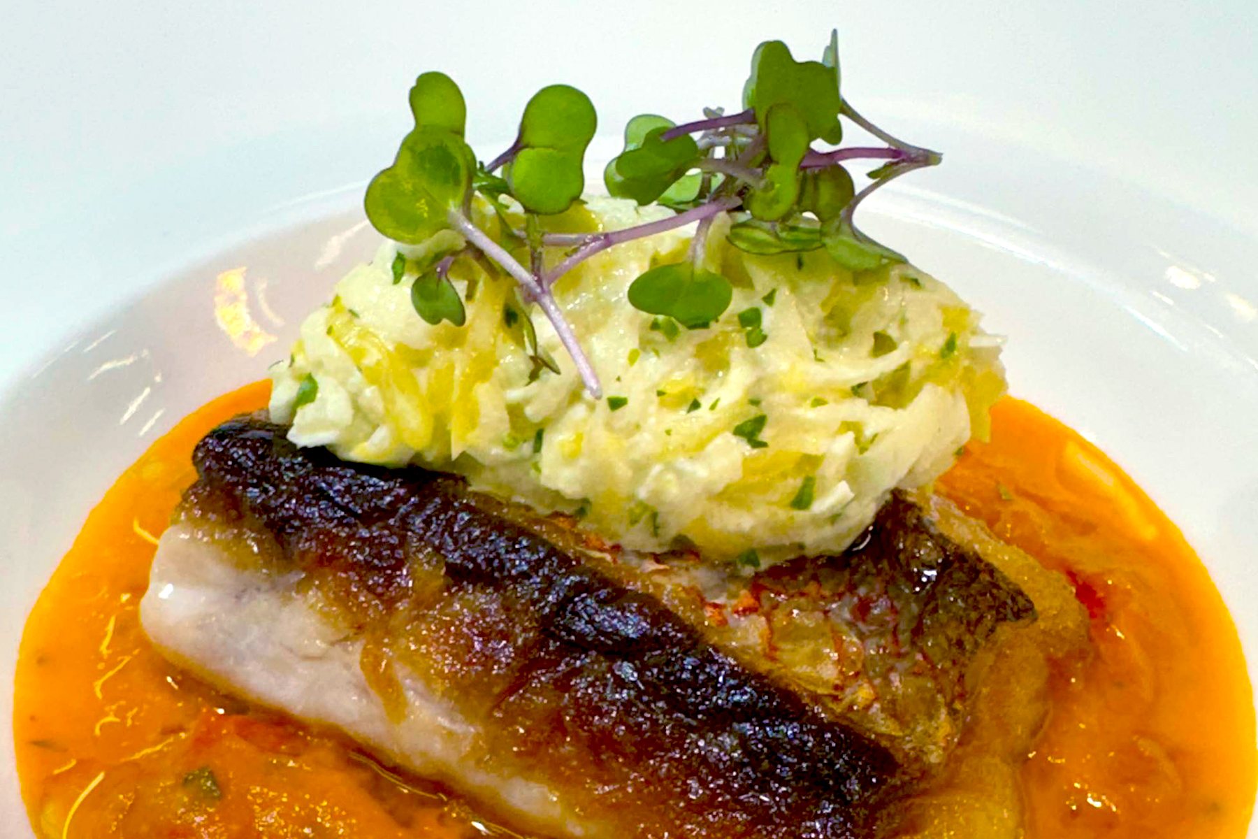 A piece of seared fish with crispy skin sits on a bed of orange sauce, topped with a creamy, shredded vegetable mixture and garnished with fresh microgreens. Fearrington Village