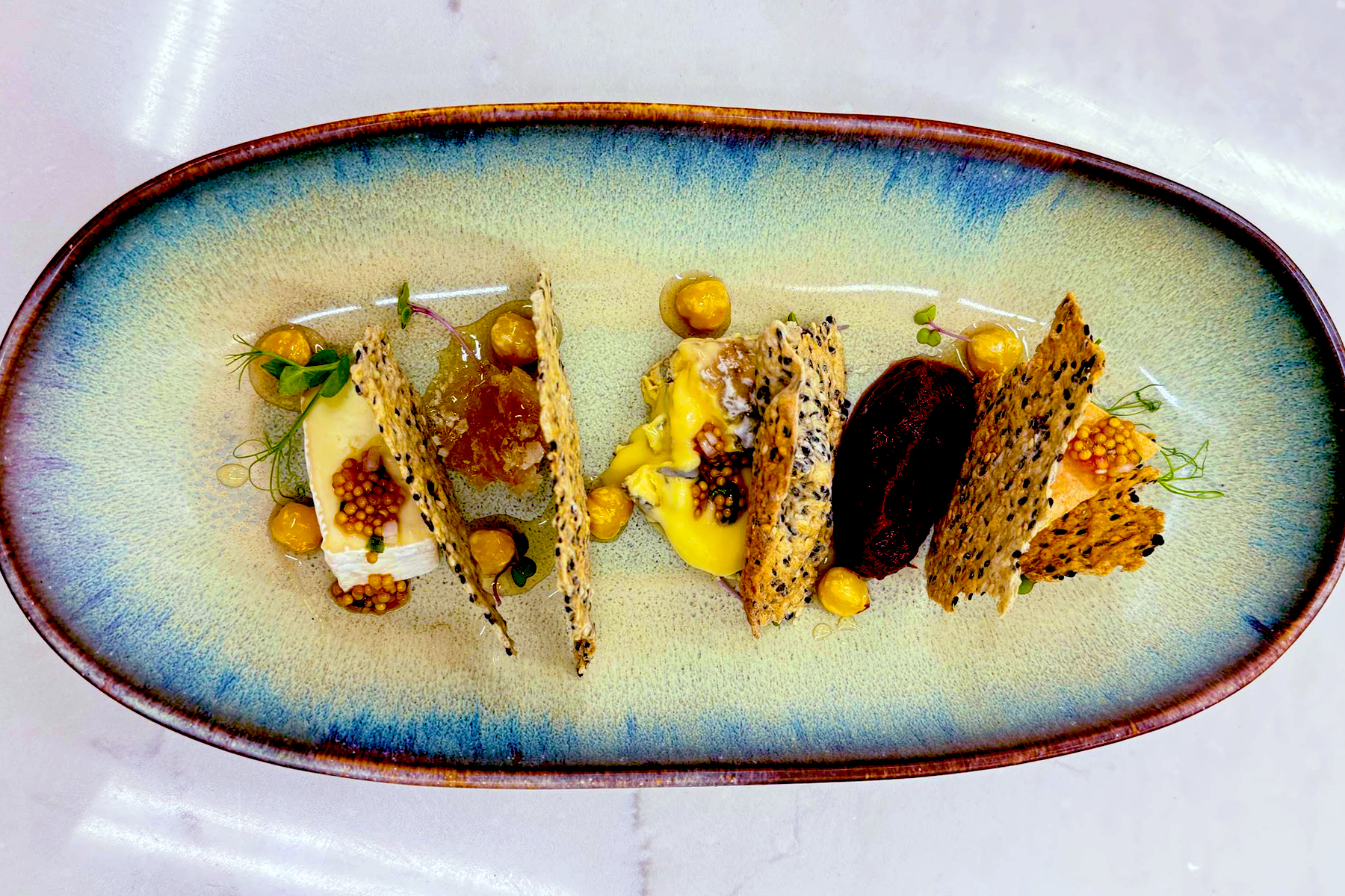 Four gourmet crackers with various toppings, including yellow cream, beet slice, herbs, seeds, and small dollops of sauce, are artfully arranged on an oval ceramic plate. Fearrington Village