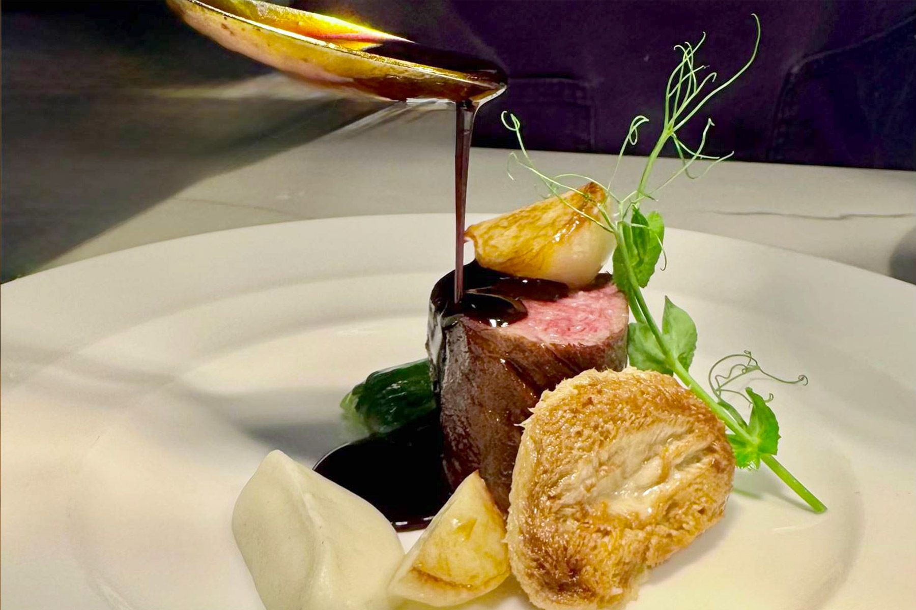 A close-up of an elegant plated dish featuring a piece of rare steak, chicken, vegetables, and a rich brown sauce being carefully poured over the meat, garnished with microgreens on a white plate. Fearrington Village