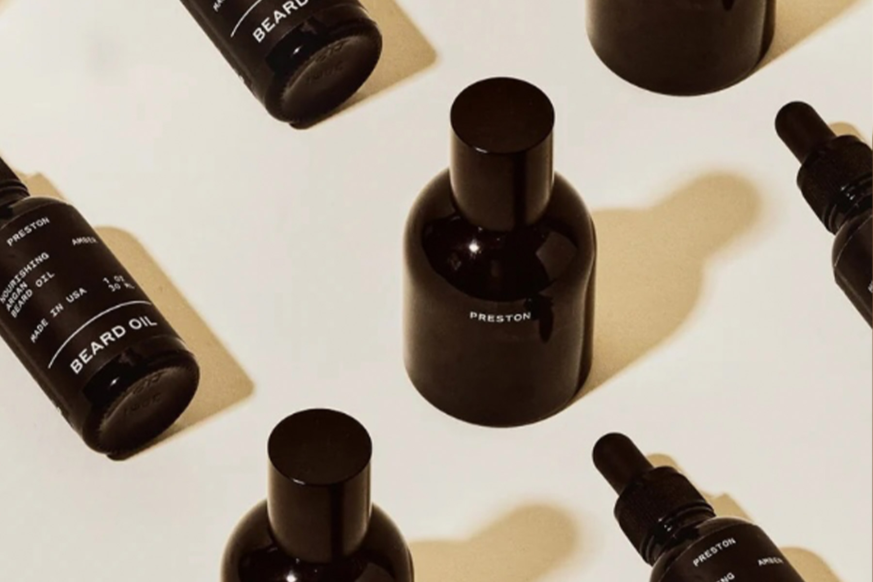Several black bottles labeled "BEARD OIL" and "PRESTON" are arranged on a beige surface, some upright and some lying flat, creating a stylish, minimalist pattern with shadows. Fearrington Village