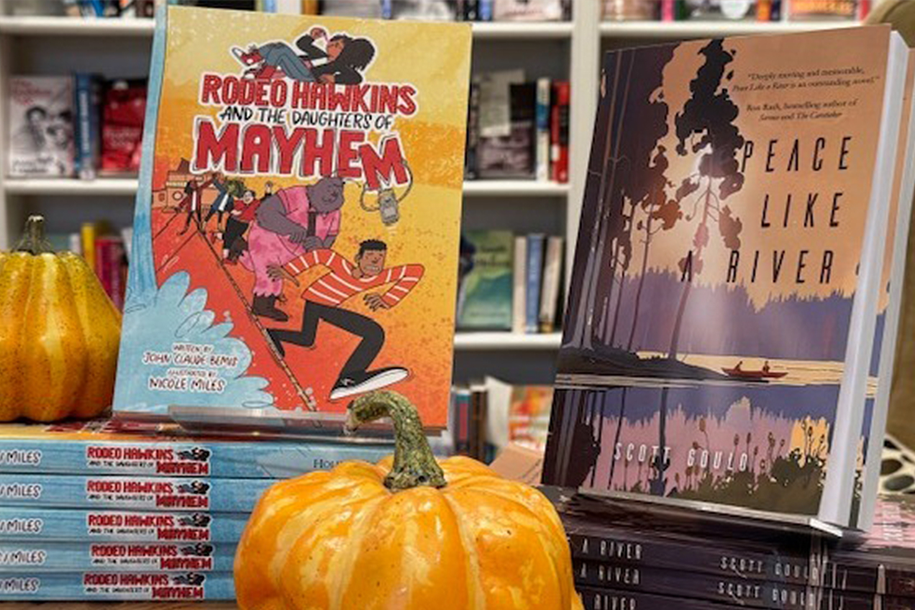 Two stacks of books are displayed on a table with small pumpkins. One book is titled "Rodeo Harkins and the Daughter's of Mayhem," featuring colorful cartoon art, and the other is "Peace Like a River," with a calm, scenic cover. Fearrington Village