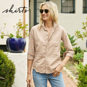 A woman wearing sunglasses, a beige button-up shirt, and light blue jeans stands outside near potted plants, smiling. The word “shirts” is written in cursive at the top left of the image. Fearrington Village