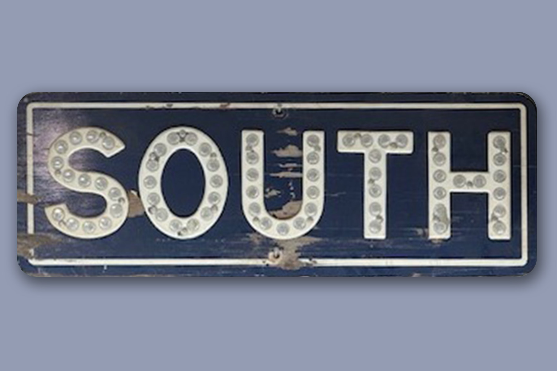 A worn blue street sign with the word "SOUTH" in large white letters, each letter containing round reflector dots. Fearrington Village