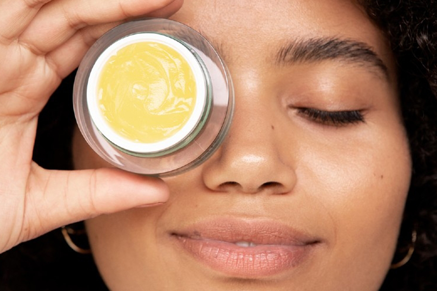 A person with closed eyes smiles softly while holding a small jar of yellow cream close to their face, near one eye. Fearrington Village