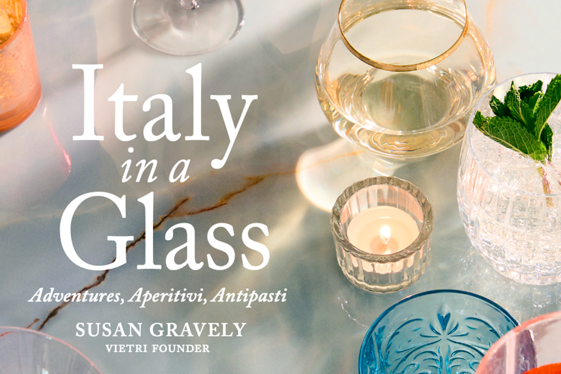 A marble table with various glasses, a candle, and a mint garnish. Text reads: "Italy in a Glass: Adventures, Aperitivi, Antipasti. Susan Gravely, Vietri Founder. Fearrington Village
