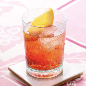 A glass filled with a reddish-brown iced drink, garnished with a lemon wedge, sits on a white tile coaster atop a pink and white patterned tablecloth. Fearrington Village