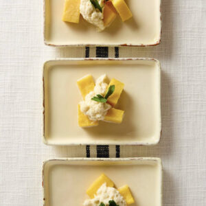 Three rectangular plates each hold a small stack of polenta cubes topped with a creamy spread and garnished with a parsley leaf, arranged vertically on a white tablecloth with a striped detail. Fearrington Village