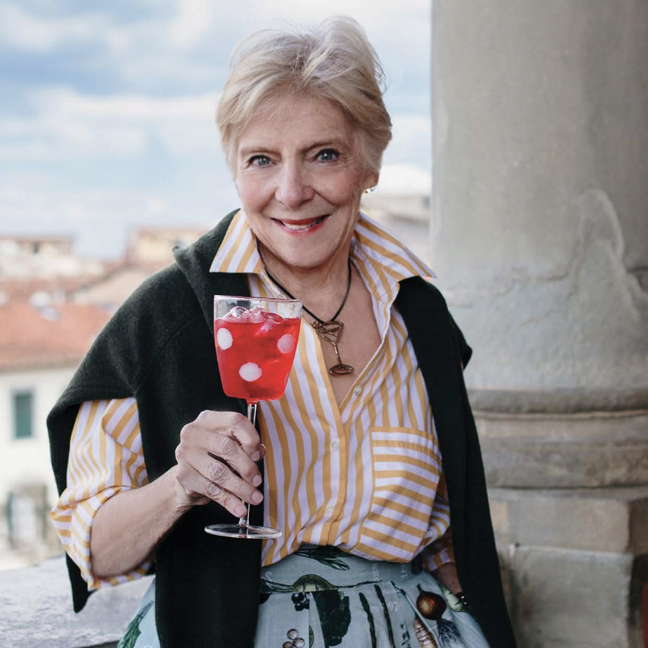 An older woman with short light hair, wearing a yellow striped shirt and a dark sweater draped over her shoulders, smiles while holding a glass of bright red drink with ice and white spots, standing outdoors by a column. Fearrington Village