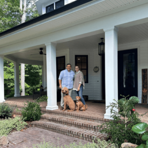 Two people stand smiling on the brick steps of a white house’s front porch, accompanied by two dogs. Lush greenery surrounds the porch, and a vertical “WELCOME” sign hangs by the dark front door. Fearrington Village