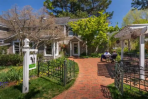 A brick pathway leads to a house surrounded by trees and greenery; two people sit at a table under a pergola on the right, and a white signpost with a horse image stands on the left. Fearrington Village