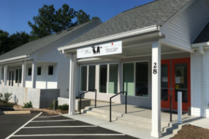 A white one-story building with a gray roof, a ramp and stairs leading to a red front door, and a sign above the entrance. The building is surrounded by a paved parking lot and some greenery. Fearrington Village