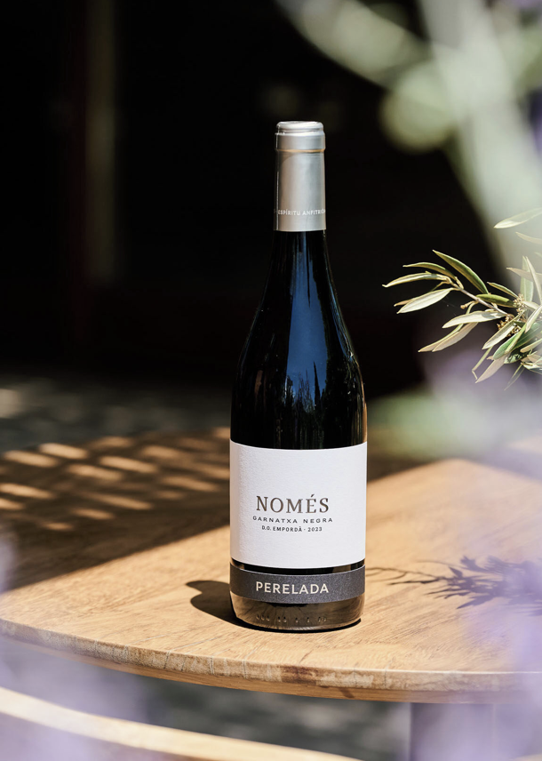 A bottle of Perelada Només Garnatxa Negra red wine sits on a wooden table, with blurred foliage and sunlight creating a relaxed, outdoor atmosphere. Fearrington Village