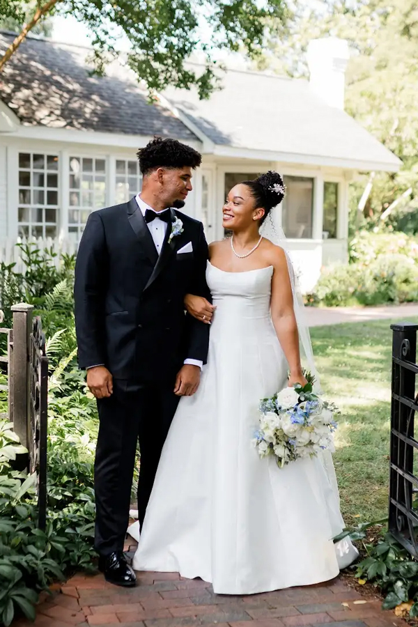 A bride and groom stand outside, smiling at each other. The bride wears a white gown and holds a bouquet, while the groom wears a black tuxedo. They are arm in arm in a garden setting near a house. Fearrington Village
