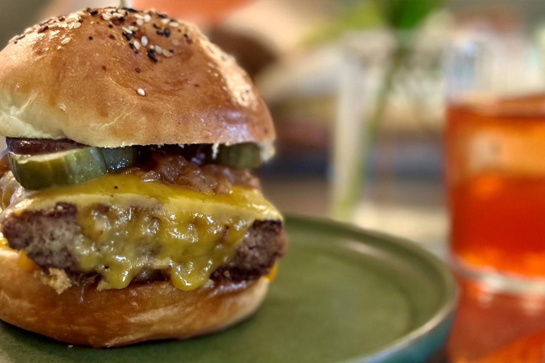 A close-up of a cheeseburger with a beef patty, melted cheddar, pickles, and onions on a seeded bun, served on a green plate. A blurred drink sits in the background. Fearrington Village