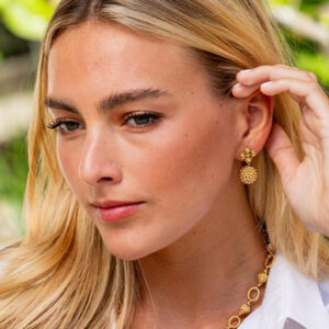 A woman with long blonde hair wears a white top, gold earrings, and a matching gold necklace, touching her ear while looking to the side with a neutral expression. Green foliage is blurred in the background. Fearrington Village