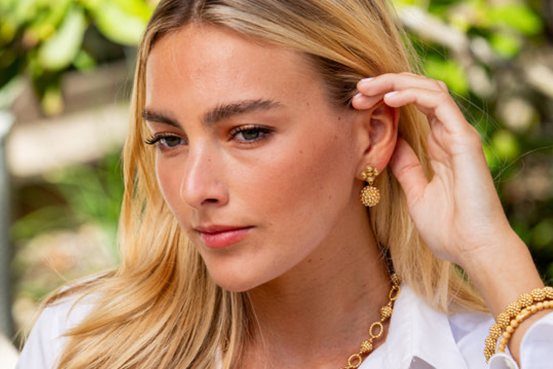 Dovecote Fall 2025 Trunk Show: Capucine de Wulf 7 A woman with long blonde hair wears a white top, gold earrings, and a matching gold necklace, touching her ear while looking to the side with a neutral expression. Green foliage is blurred in the background. Fearrington Village