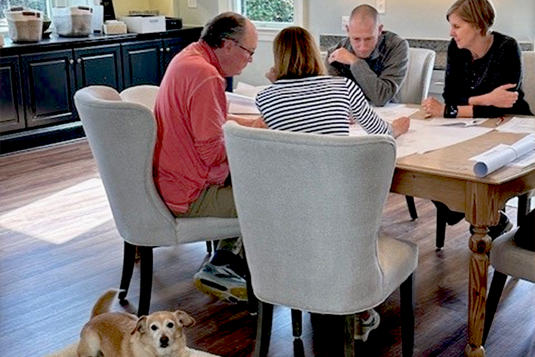 Four adults sit around a dining table looking at papers, appearing to discuss or review something. A small brown and white dog lies on a rug in the foreground, looking toward the camera. Fearrington Village