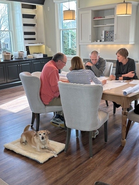 Four adults sit around a dining table looking at papers in a bright room with large windows; a small brown and white dog lies on a mat on the floor nearby, looking toward the camera. Fearrington Village