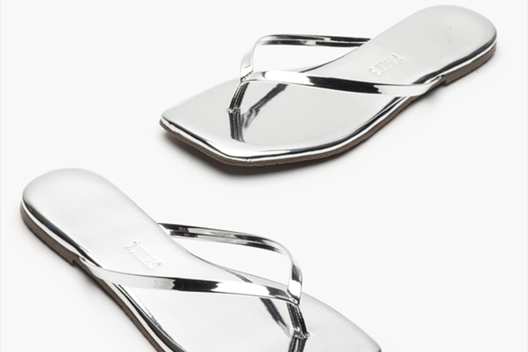 A pair of shiny, metallic silver flip-flop sandals with thin straps and slightly square toes, displayed on a plain white background. Fearrington Village